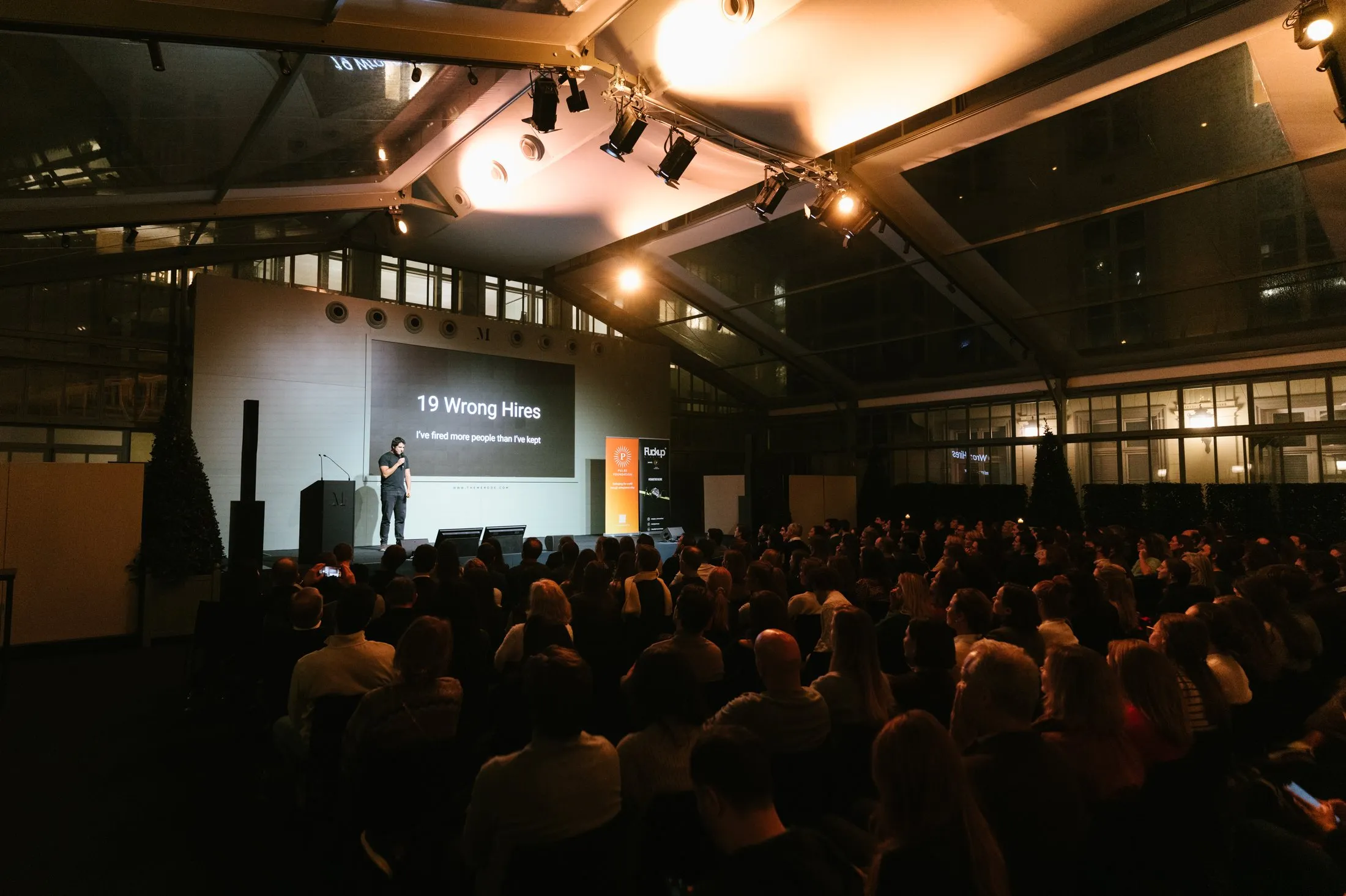 Une salle sombre sous une verrière, un public dense, et un entrepreneur sur scène à côté d'une diapositive qui annonce dix-neuf mauvais recrutements.